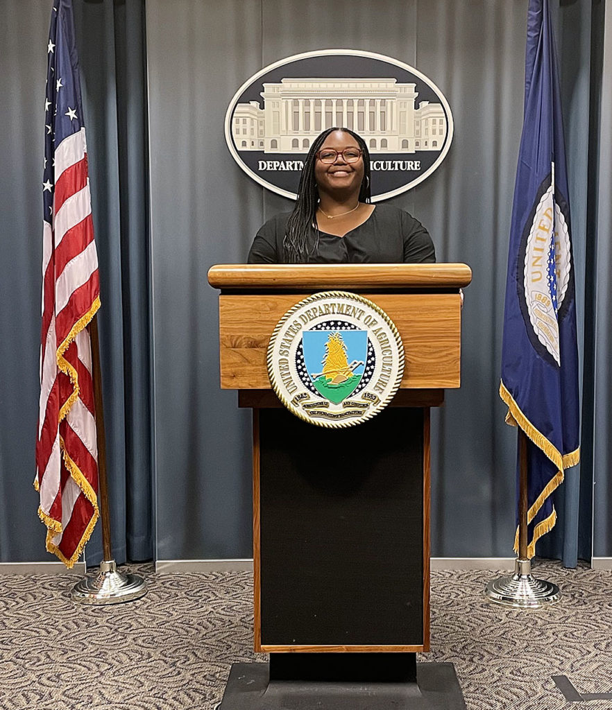 a woman, Mickeala Carter, standing at podium at USDA with flags on both sides of her