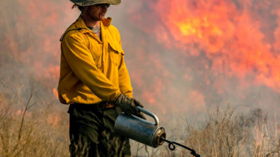 A man wearing a yellow shirt and brown hat uses a tank to ignite dry brush for a prescribed burn. There are flames behind him,