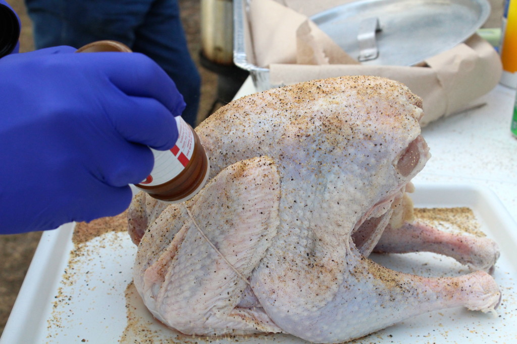 Blue-gloved hand sprinkles seasoning salt on a raw turkey being dry brined for cooking. 