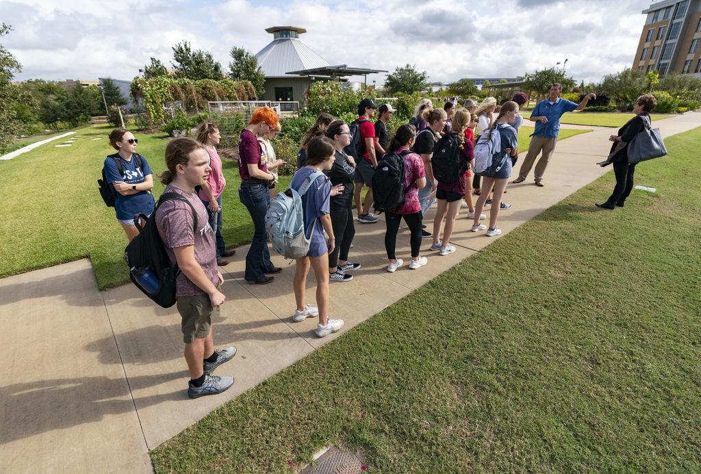 A group of Texas A&M University students stand on a sidewalk in the Gardens with buildings in the background