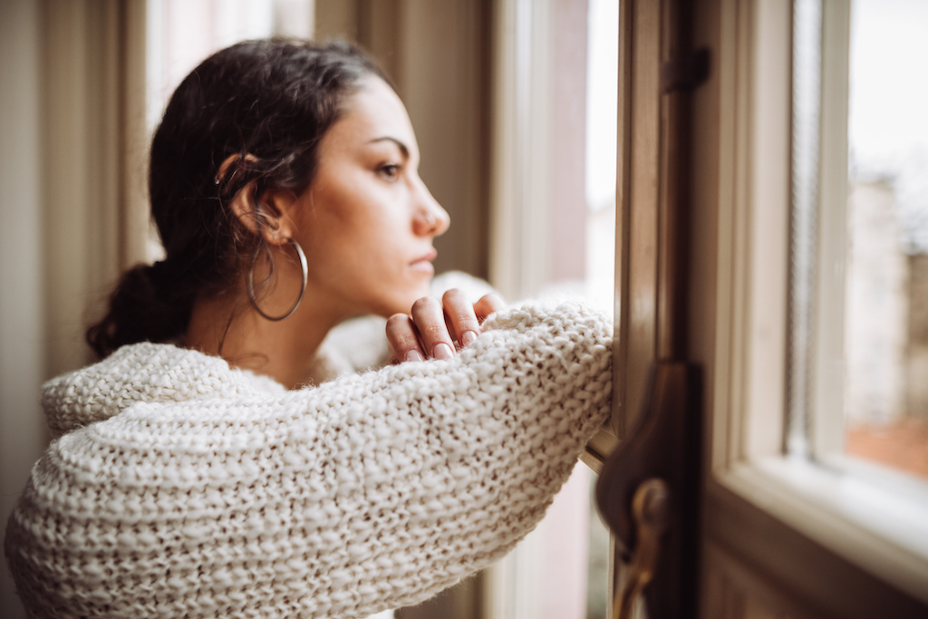 A woman gazes pensively out a row of windows. Her dark hair is in a low ponytail and she wear a beige baggy sweater. 