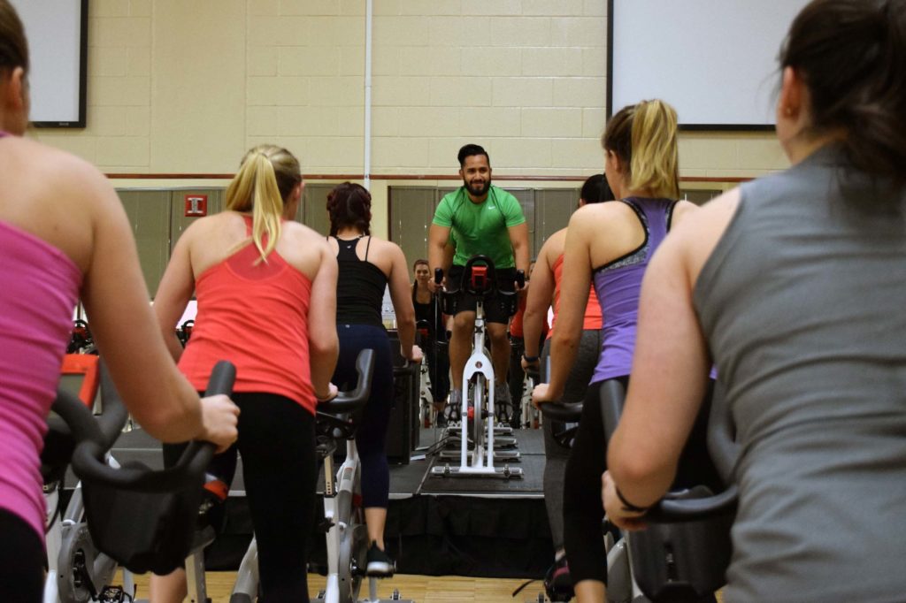 A spin cycle class with a man in a green shirt at the front of the room facing the camera