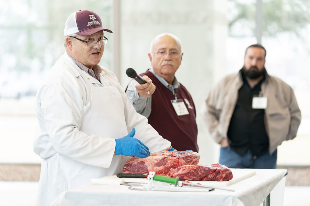 Three men stand at the front of a room, one of them in front of a pile of raw beef and he is holding a microphone.