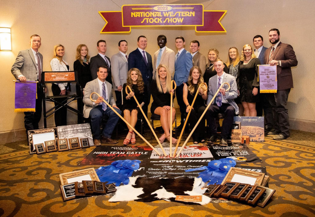 a large group of college-age students holding show canes and banner and a wide range of awards spread out on the floor in front of them.