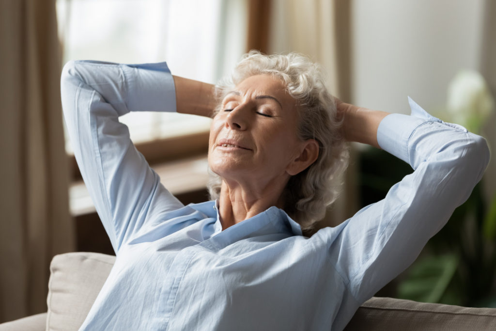 An older woman indulges in mindfulness by leaning back in a chair with eyes closed and resting in her hands behind her head.