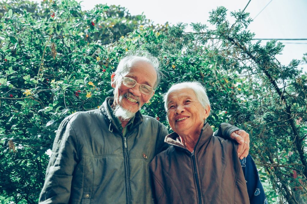An elderly couple, one man and one woman, in dark windbreakers stand smiling in front of a green tree. Photo is from the waist up.