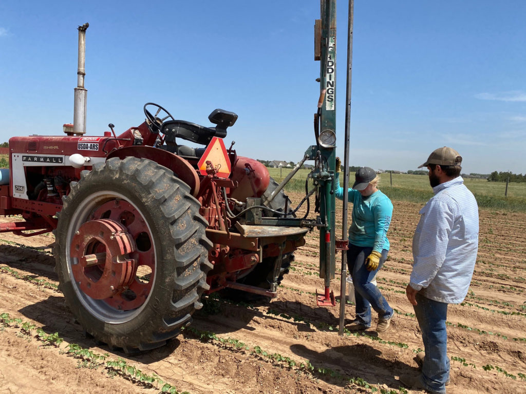 two students stand with a tractor as a coring machine takes a deep soil test to measure fertility