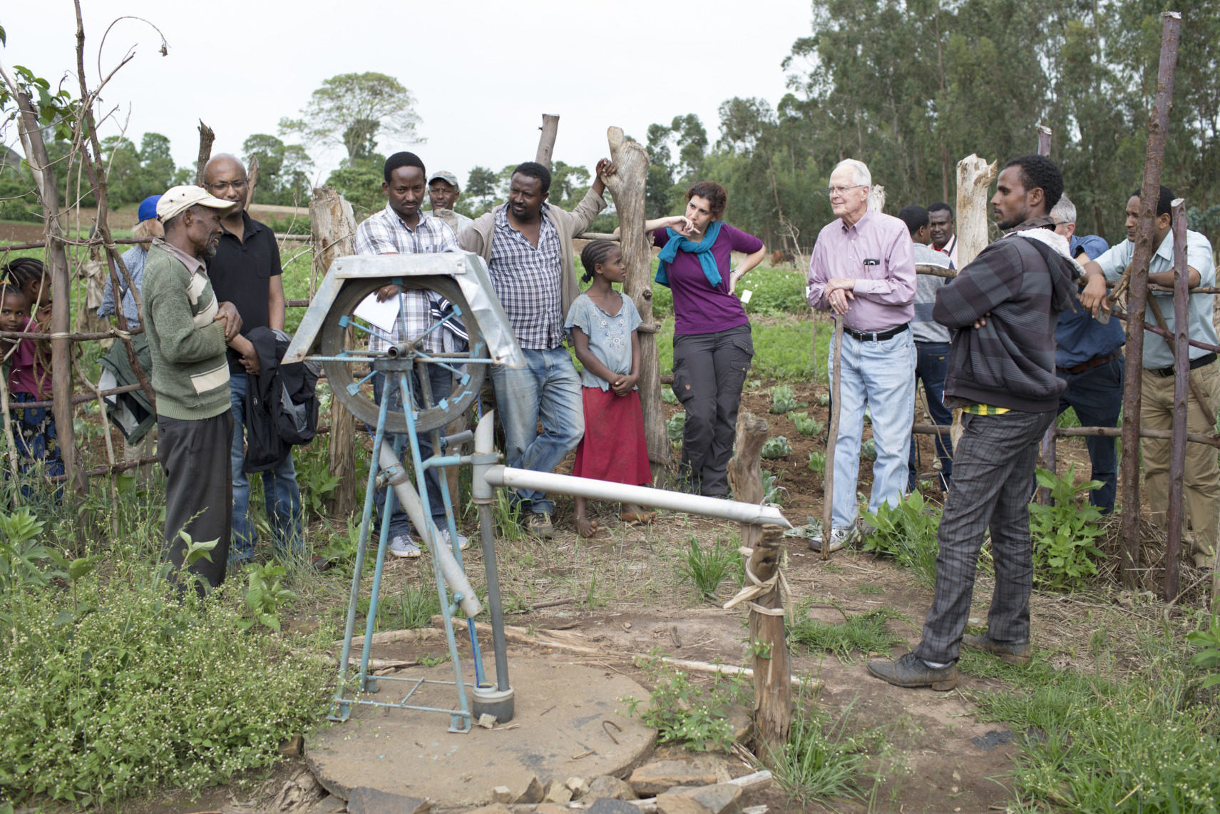 A group of people, both scientists and families, stand outside around a water pump in a jungle-looking area.