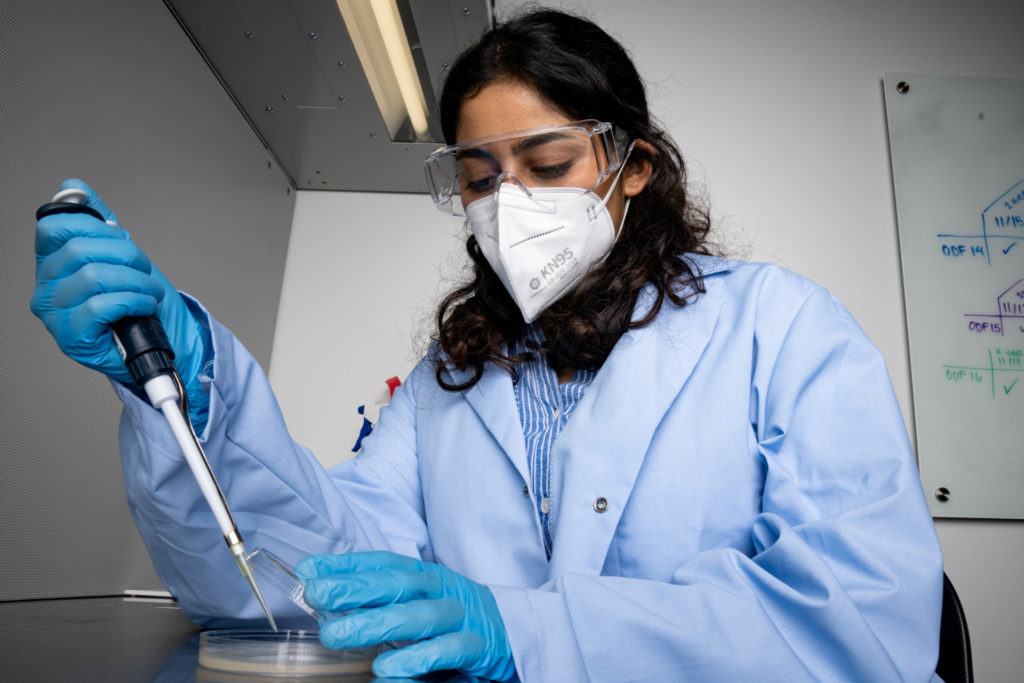 Female student in mask, goggles, and lab coat uses pipette to fill petri dish.