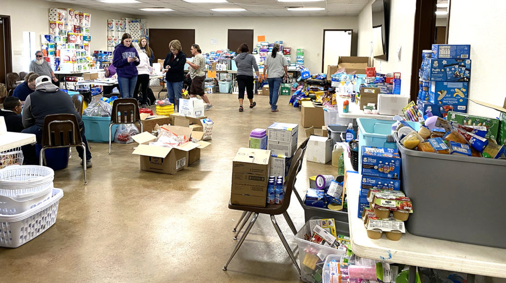 A large room full of tables and chairs stacked with diapers and toiletries and other donated items with people working in the back to distribute to people who lost during the tornadoes