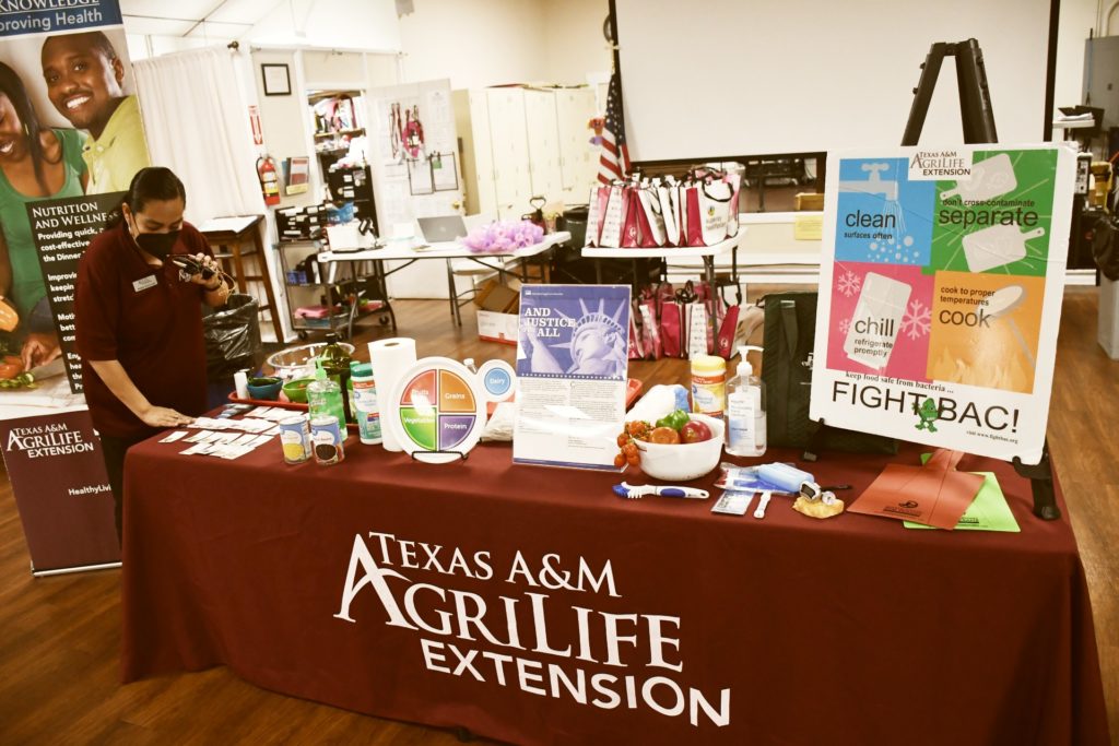 A lady, Angie Gutierrez, stands beside a table full of information and posters as she calls to participants at nutrition bingo educational program.