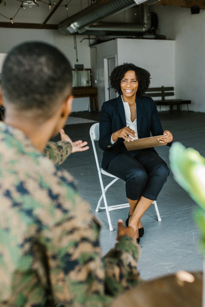 A woman in a business suit sits on a chair. She is talking to a military member, wearing camouflage, who is also in a chair but facing away from the camera. 