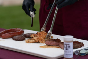 Various meat being cut after grilling with a Texas A&M branded spice bottle in front. 