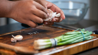 A pair of hands peel a garlic bulb. Under their hands in a cutting board with a bunch of green onions.