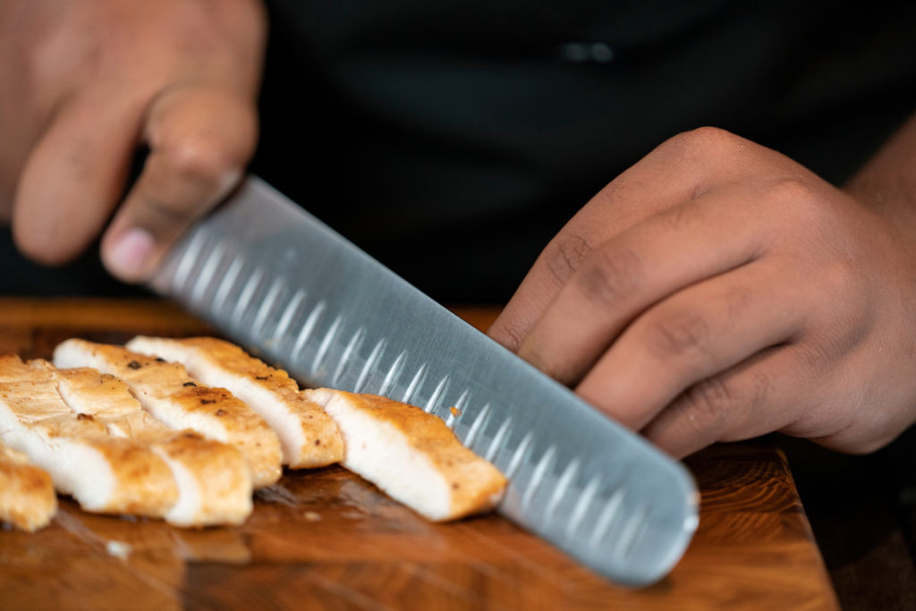 A close up of a pair of hands holding a knife. On the wood table is a cooked chicken breast that is being cut in large slices.