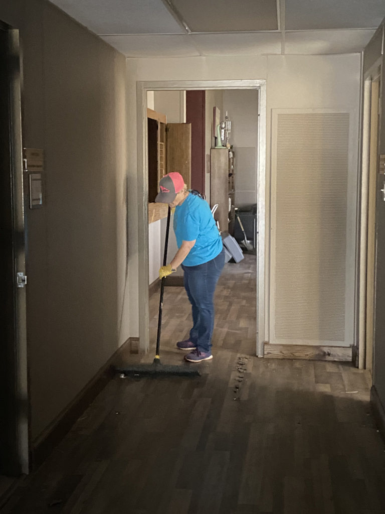 A woman in a blue t-shirt sweeps in a dark hallway of the damaged building.