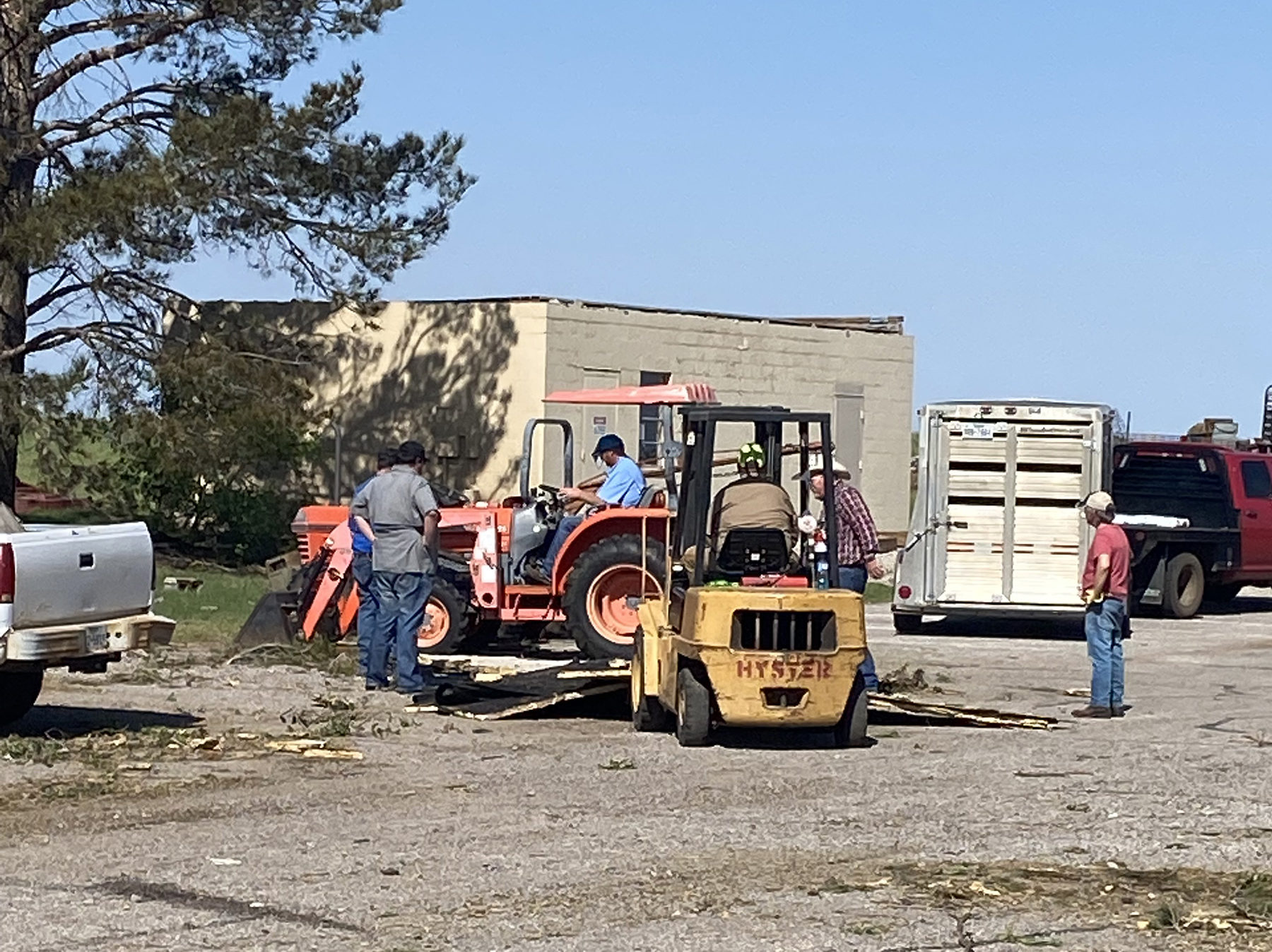 Five men are standing and sitting around a tractor and skid loader as debris is being picked up and loaded into a trailer.