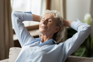 Older woman leaned back, eyes closed, arms reaching back and hands behind the head, relaxing