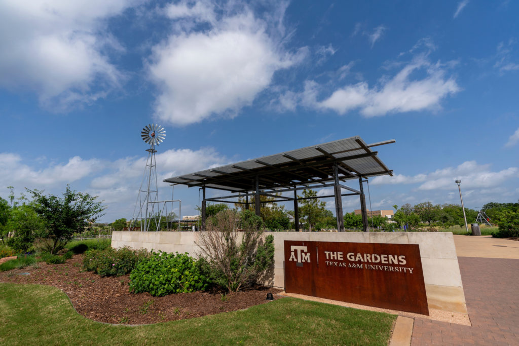 An entry sign that says The Gardens Texas A&M University with greenery in front and a bricked path to the side