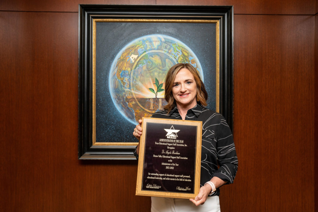 A woman stands holding a plaque in front of a framed Texas A&M University symbol.