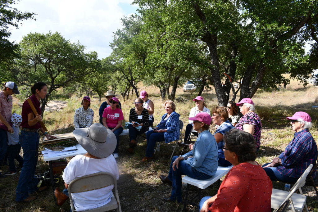 A group of ladies sitting outdoors, many with pink hats on, and listening to a presentation.