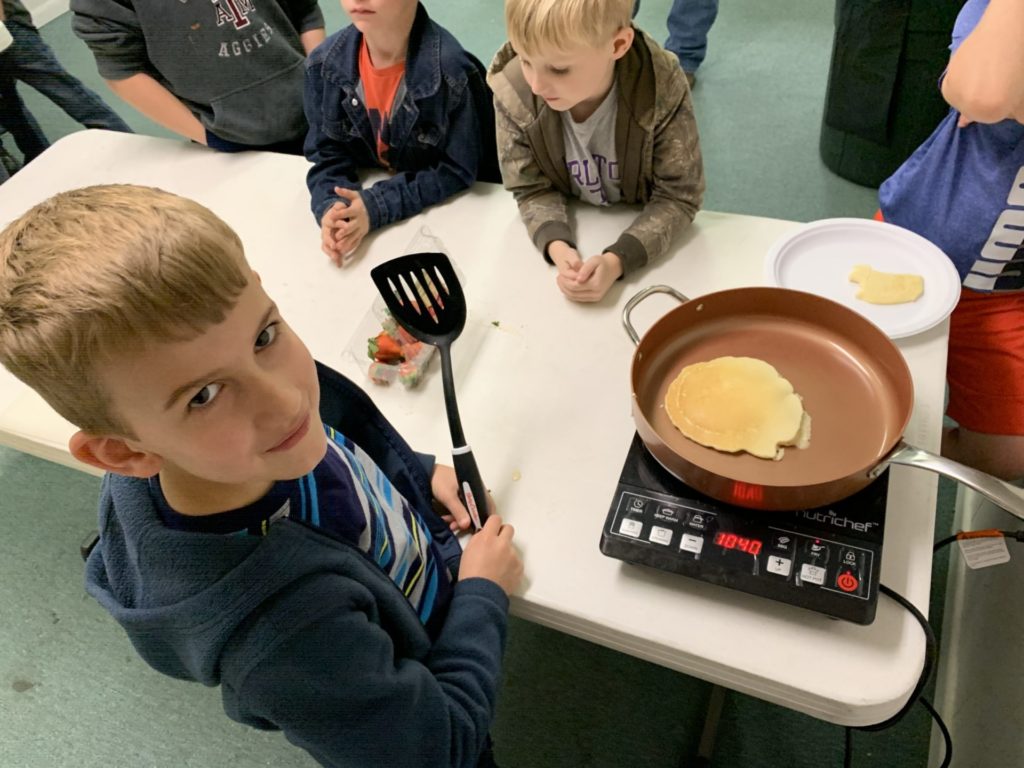 A boy is looking up at the camera and beside him is a pancake cooking in a copper skillet on a hot plate with other children looking on during camp.