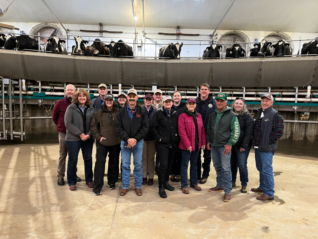 A group of people stand in the middle of a dairy parlor with dairy cattle looking on above.