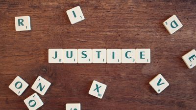 Scrabble word game tiles spell out the word justice. They set on a wood table with randome other letter titles scattered around them