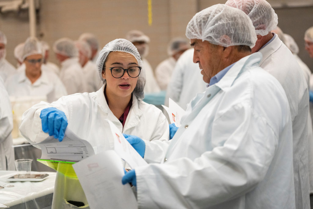 a woman and a man in hair nets, gloves and lab coats discuss paperwork in a barbecue class.