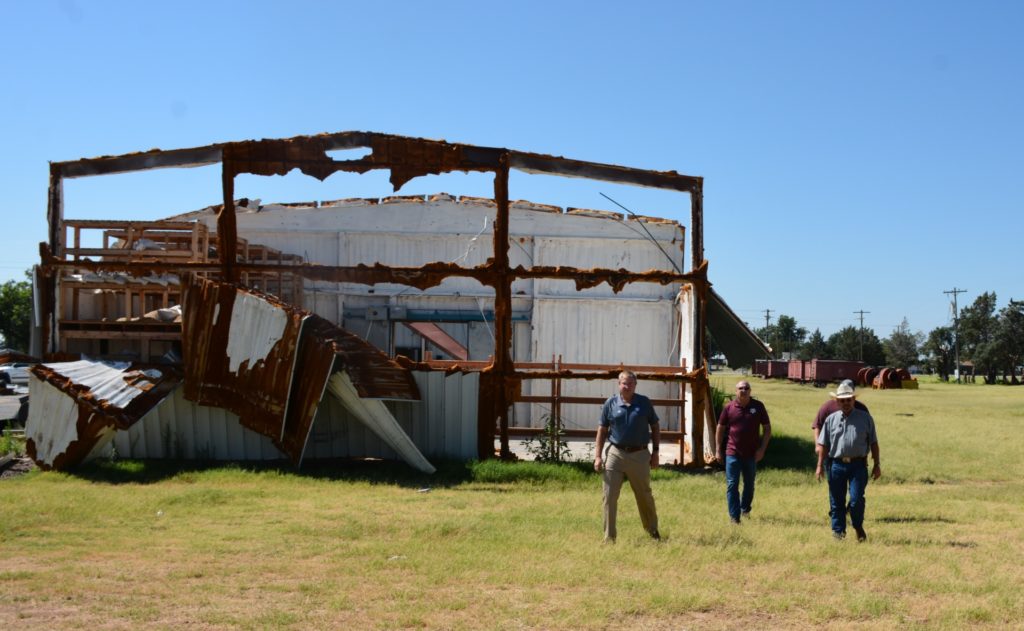 The skeleton steel beams of a barn can be seen after a tornado ripped the sheet metal off the front and sides. Four men are walking in front of it.