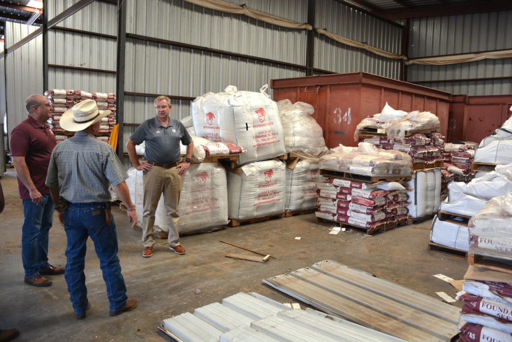 Three men stand in a warehouse that has it's walls lined with large white totes of seed and bags of seed on pallets.