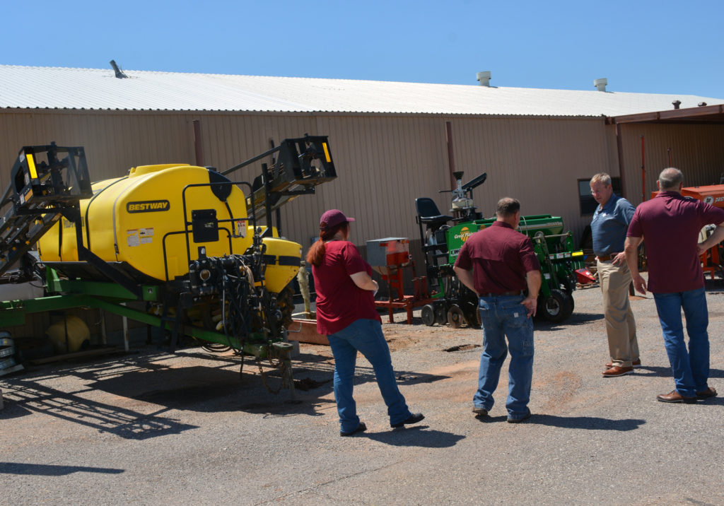 a large yellow tank on a sprayer and a piece of green equipment can be seen behind a group of people viewing the damage.