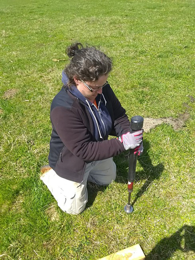 A woman is down on her knees in the grass holding onto a black piece of equipment that is taking measurements to determine sustainability of practices
