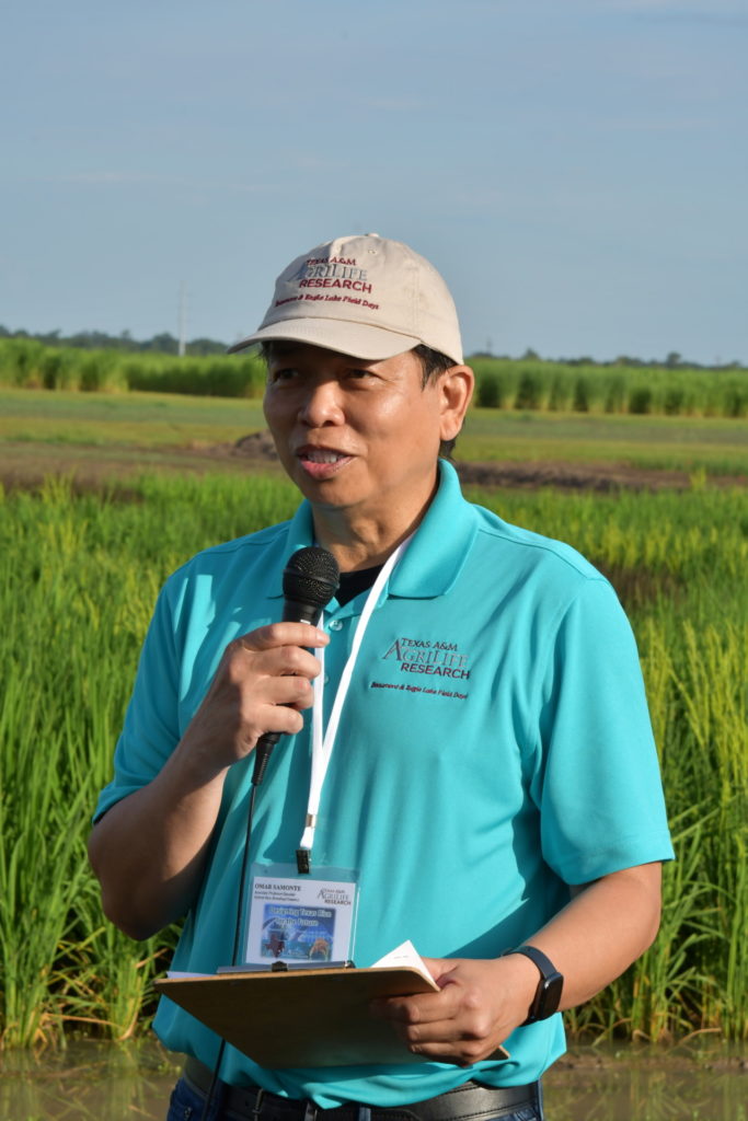 A man in a turquoise shirt talks on a microphone in a green rice field.
