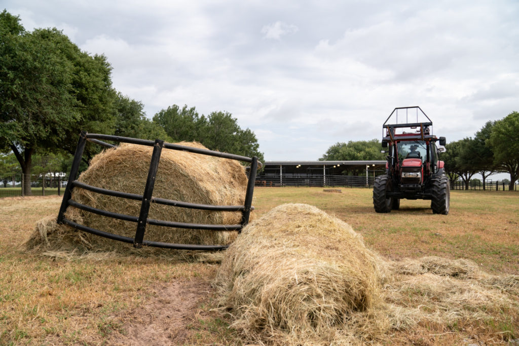 A round bale of hay is piled on the ground and in a round pen with a tractor driving away