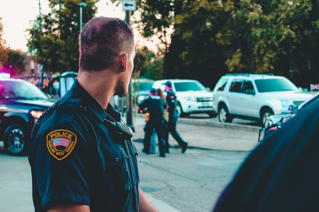 A law enforcement officer looks over his shoulder at other police officers on the street. He has close-shaven hair, a black uniform with his police patch on teh shoulder, His face is not visible.