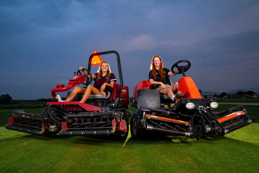 Two female students who were able to join a Little League grounds crew sit on large lawn mowers on a turf field in the early morning hours