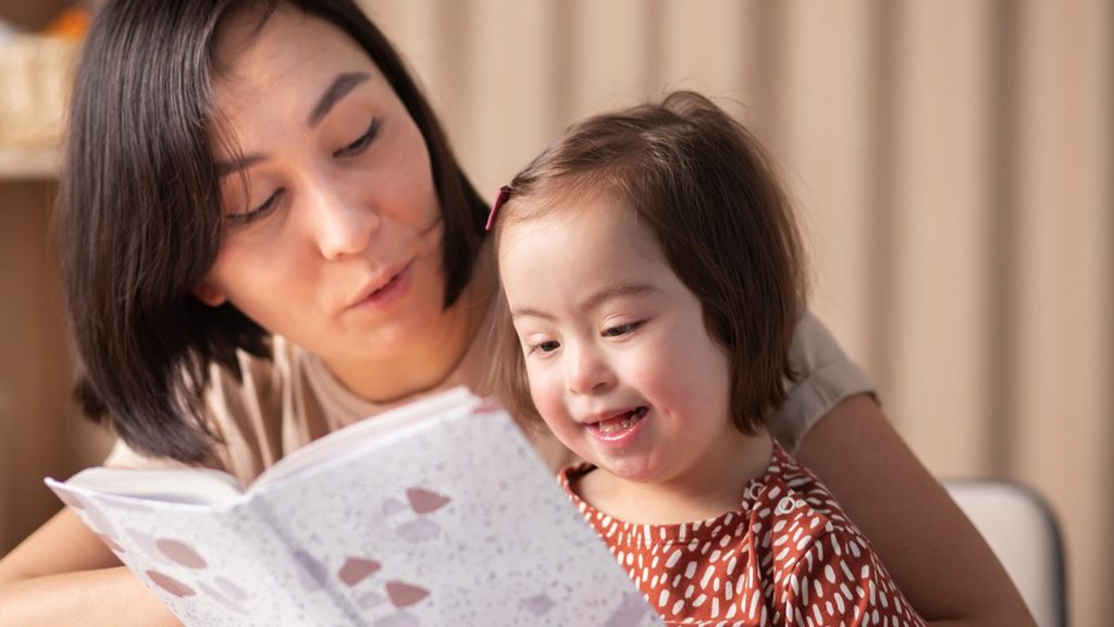 A mother reads from a storybook while her young daughter sits on her lap.