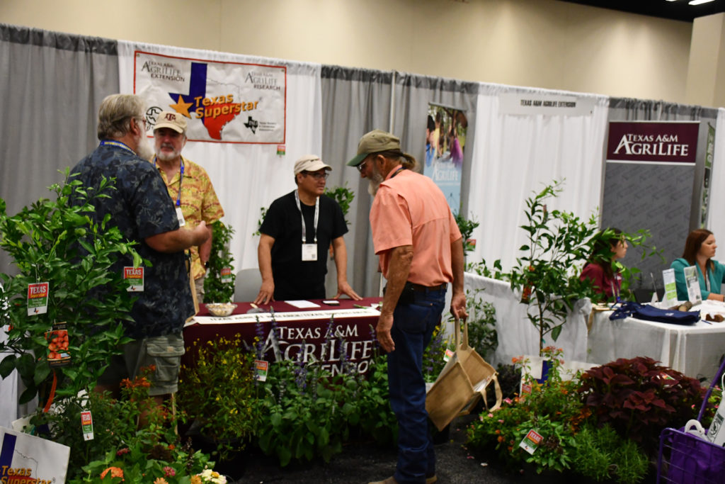 People stand on both sides of a table at a booth with greenery all around and a Texas Superstars banner up top and Texas A&M AgriLife banner below