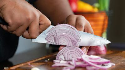 A woman's hands holding a knife cutting a purple onion in thin slices on a cutting board