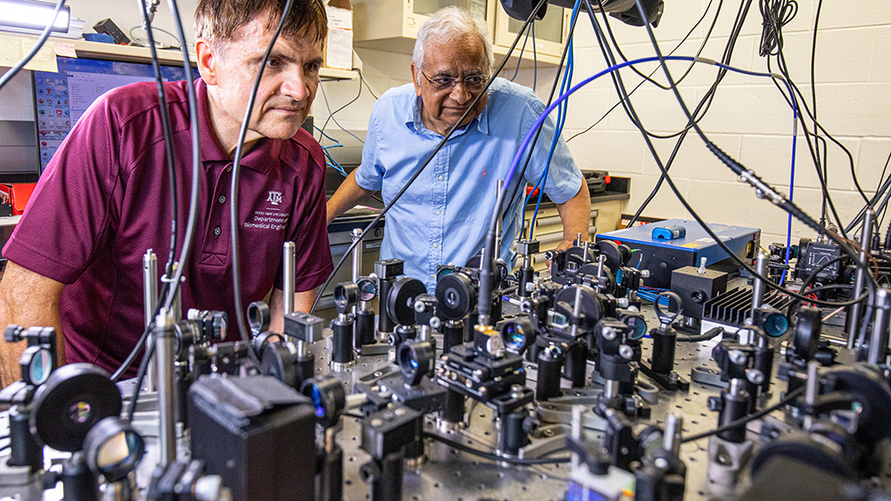 Dr. Vladislav Yakovlev and Dr. Girish Agarwal inspect a large metal table surface filled with numerous positioned devices that contain circular glass lenses and other light-focusing optics