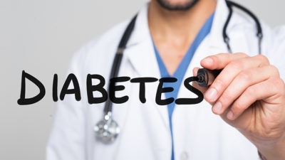 A doctor in a white coat with stethoscope writes the word Diabetes on a clear board facing the camera