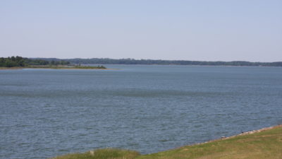 Possum Kingdom Lake. A green shore is in the foreground and the blue lake stretches almost to the horizon. There is a slight chop to it and no watercraft on it.