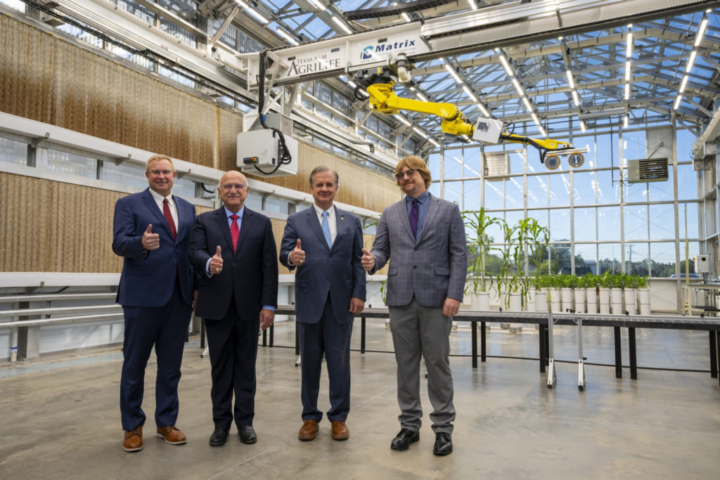 Four men showing the Gig'Em sign - thumbs up - surrounded by the Automated Greenhouse Facility
