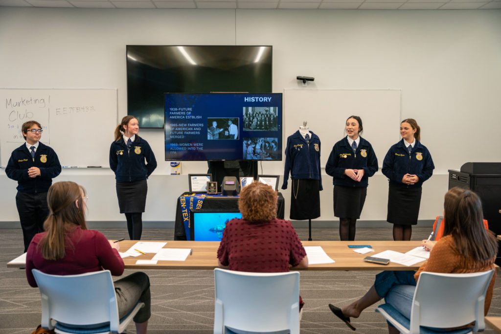 A group of students dressed in the blue FFA jackets stand before three judges seated at a table.