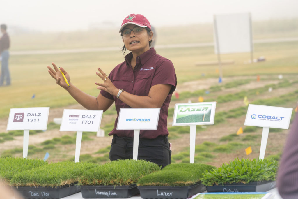 a woman, Ambika Chandra, stands in front a five different lines of turfgrass.