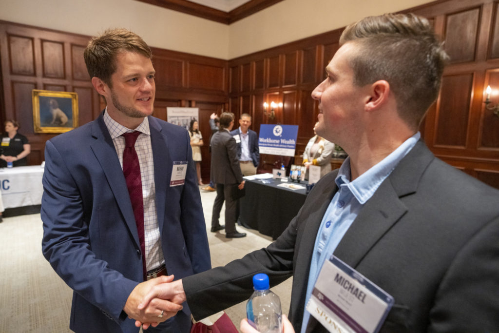 Two men, one facing forward, Nathan Harness, Ph.D., shake hands at the financial planning conference with booths behind them.