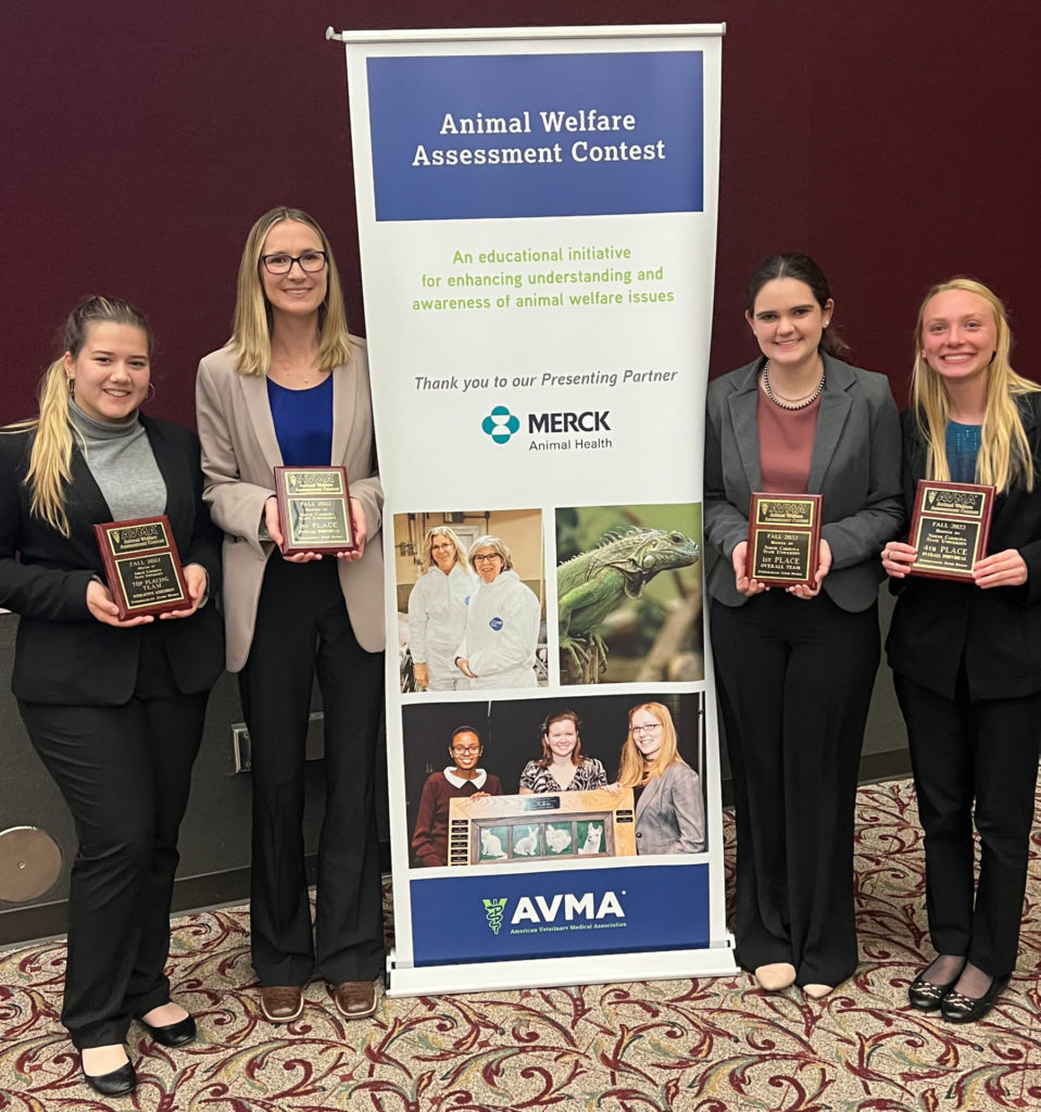 Four young women, two on each side of a large banner advertising the Animal Welfare Assessment Contest and sponsorship by Merck and AVMA, hold plaques to indicate their win.