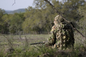 Hunter in camouflage kneels in brush looking out over an open field.