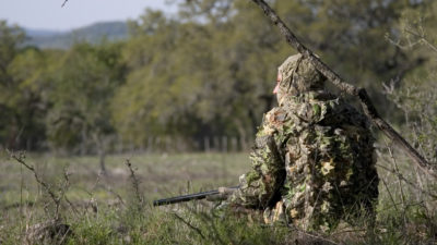 Hunter in camouflage kneels in brush.